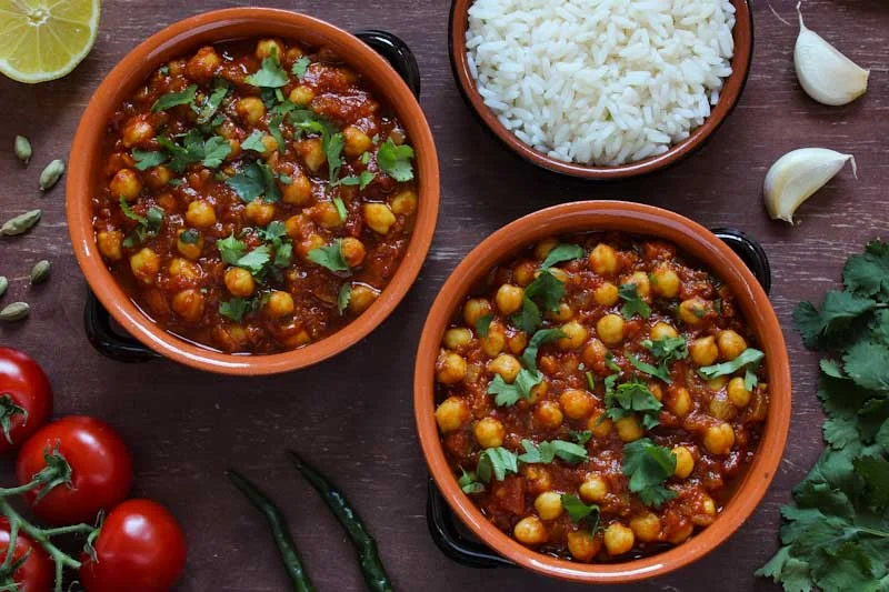Two bowls of chickpea curry garnished with fresh herbs, served alongside white rice, garlic, and lime. Fresh vegetables enhance the presentation.