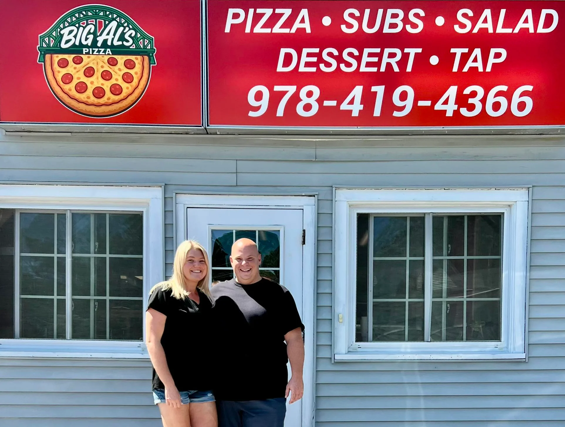 Two people standing and smiling in front of the Big Al’s Pizza shop, beneath a red sign advertising pizza, subs, salad, dessert, and tap with a phone number.