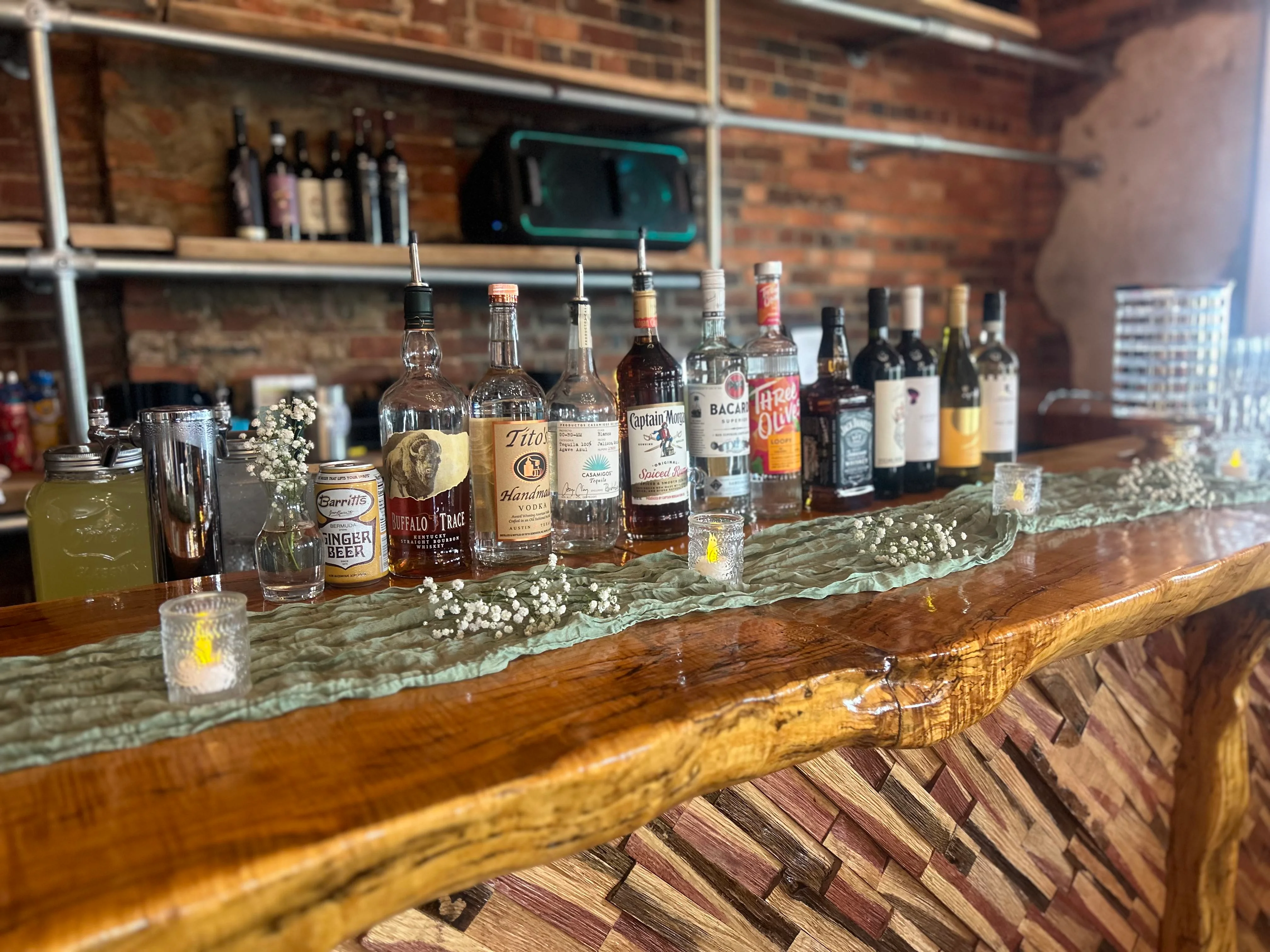 A bar counter displaying a variety of alcoholic beverages including spirits, mixers, and garnishes. The setting features wooden textures and decorative elements.
