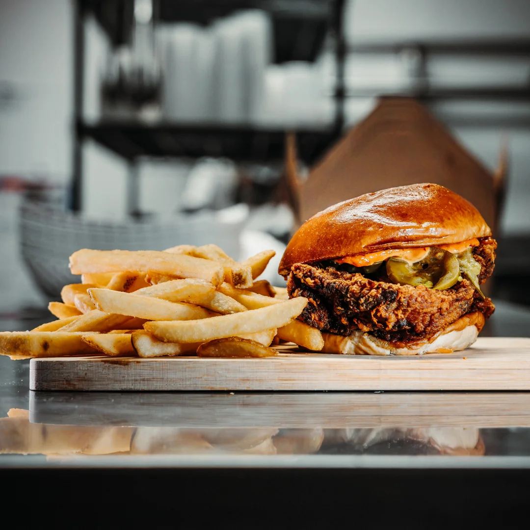 A crispy fried chicken sandwich topped with pickles and sauce, served alongside golden French fries on a wooden board.