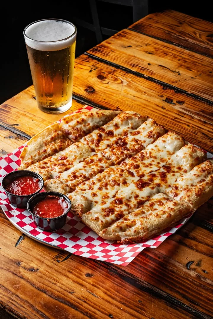 A plate of cheesy breadsticks served with marinara sauce and a glass of beer on a wooden table.