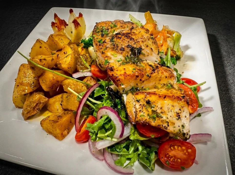 A serving of grilled fish fillet on a bed of mixed greens, cherry tomatoes, and red onions, accompanied by roasted golden potatoes.