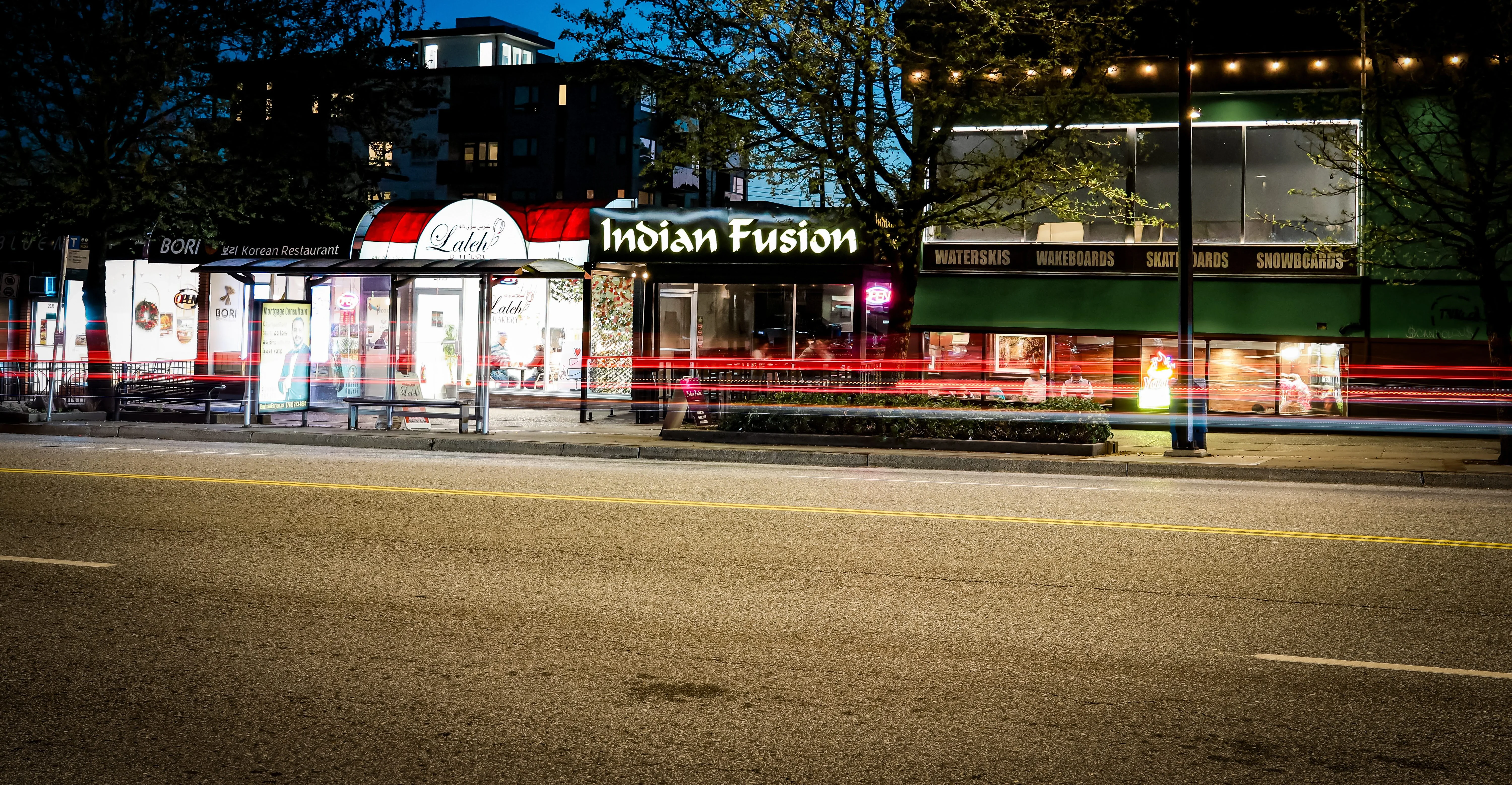 A city street at dusk featuring a variety of restaurants. One restaurant, Indian Fusion, is prominently lit with signage, while another, Laleh, is also visible. The scene includes street trees and passing car lights.