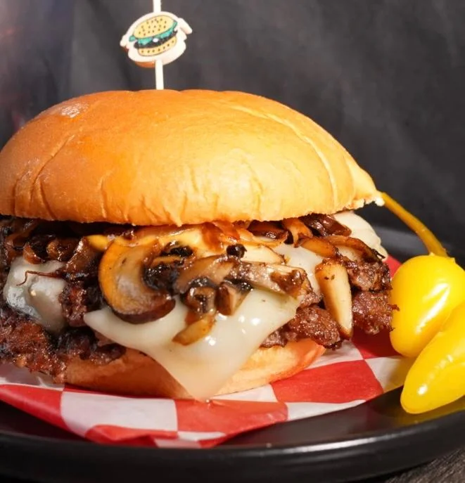 A mouth-watering burger featuring melted mozzarella cheese, grilled mushrooms, and a juicy beef patty, presented with a decorative skewer and accompanied by pepperoncini.