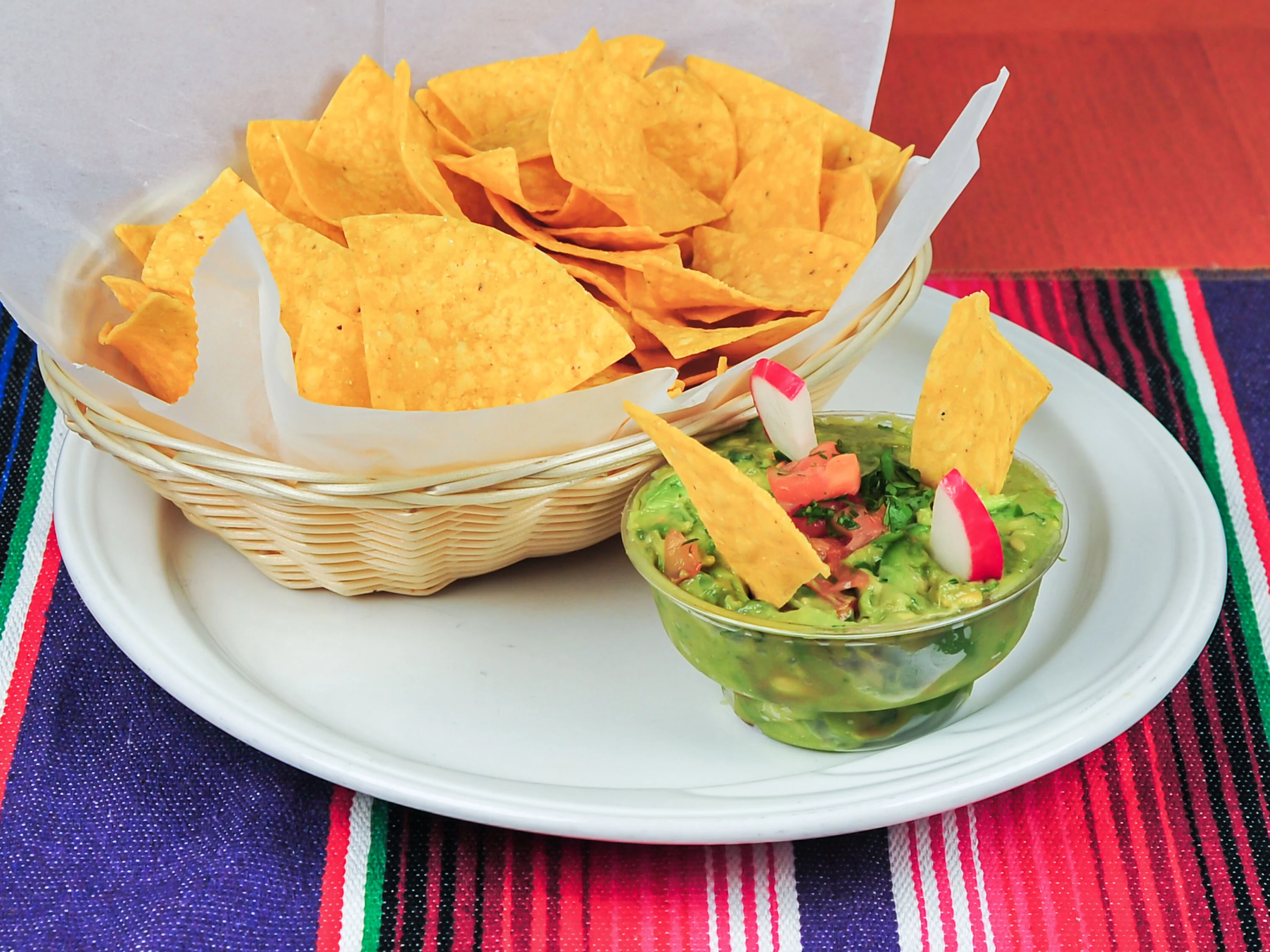 A basket of crispy tortilla chips served alongside a bowl of guacamole garnished with fresh ingredients. The presentation is vibrant against the colorful table runner.
