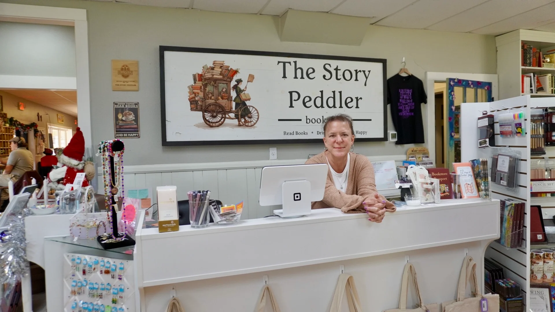 A cozy bookstore checkout area featuring a smiling attendant. The space is adorned with various books and merchandise, creating a warm atmosphere.