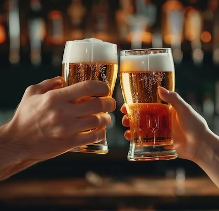 Two hands clinking beer glasses in a lively bar setting.