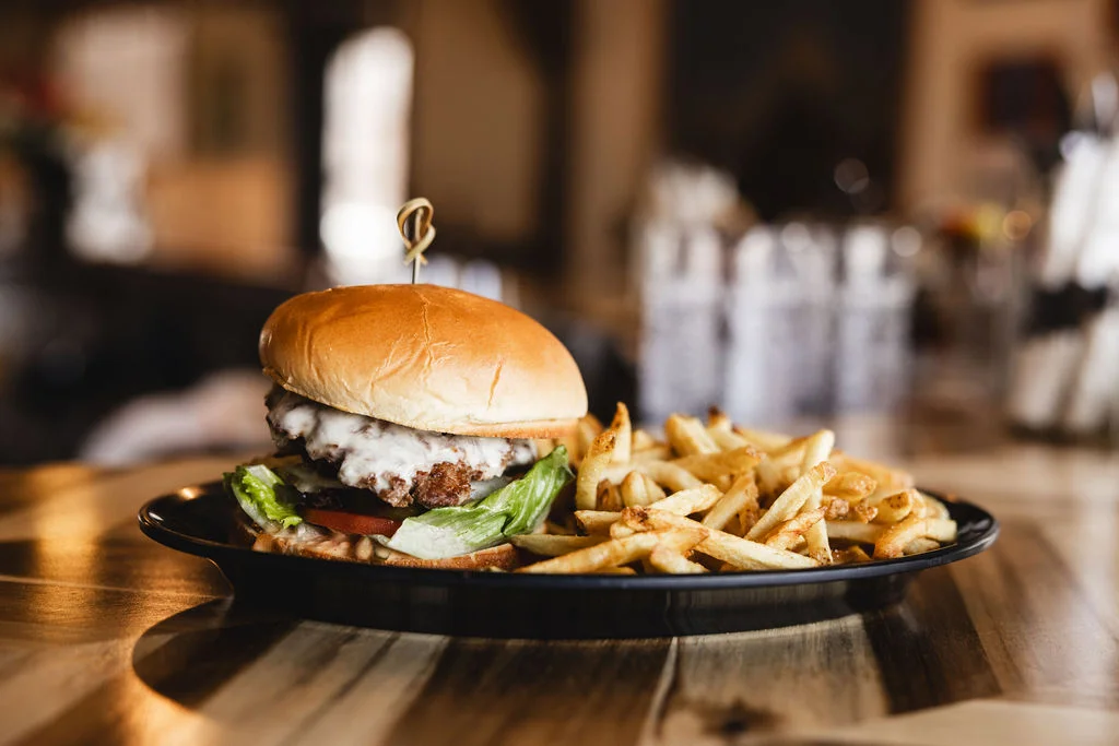 A gourmet hamburger served on a black plate with crispy French fries. The burger features layers of lettuce, tomato, and a creamy sauce, with a golden bun on top.