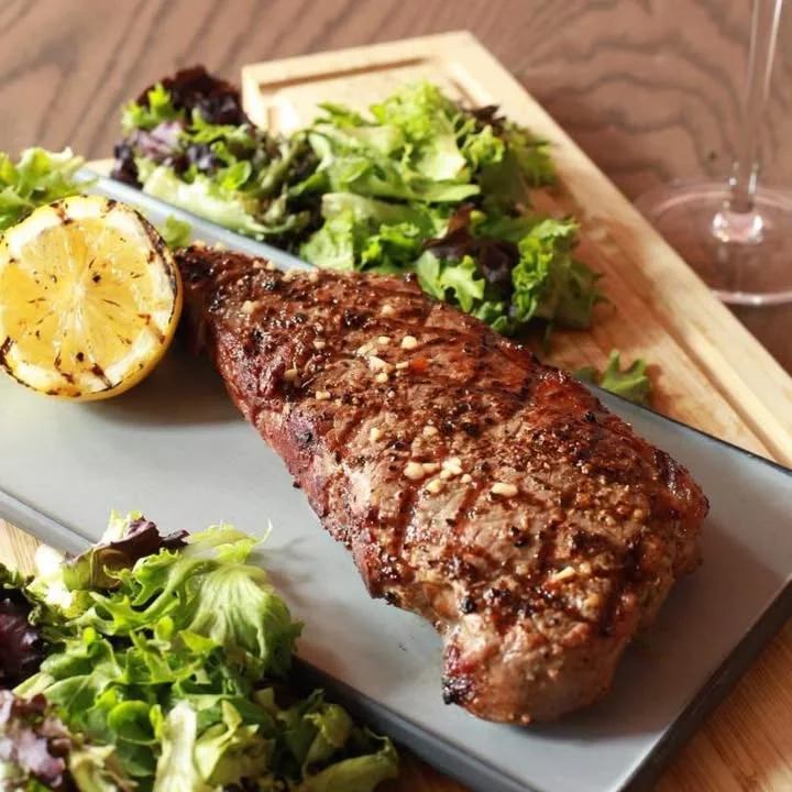 A grilled steak garnished with herbs, accompanied by a slice of lemon and fresh greens on a gray plate.
