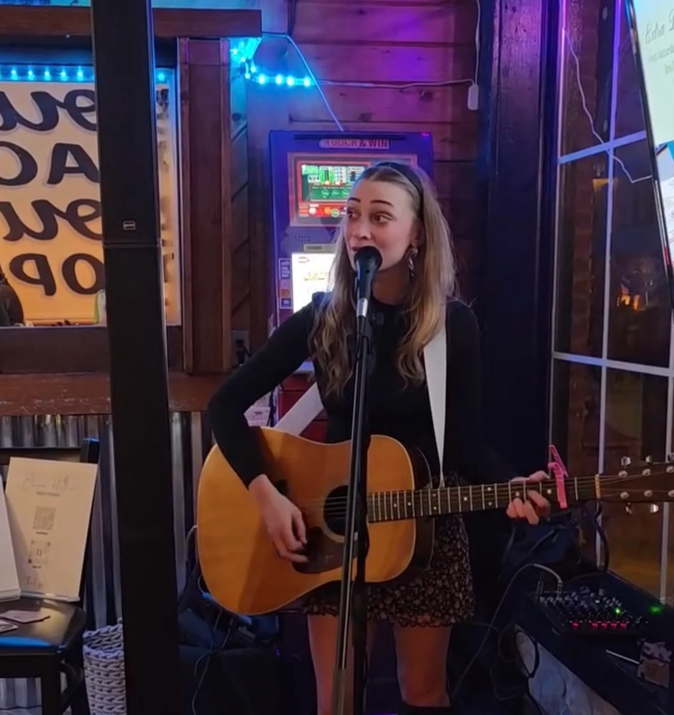 A young woman performs on stage with a guitar in a cozy interior, featuring ambient lighting and a lively atmosphere.