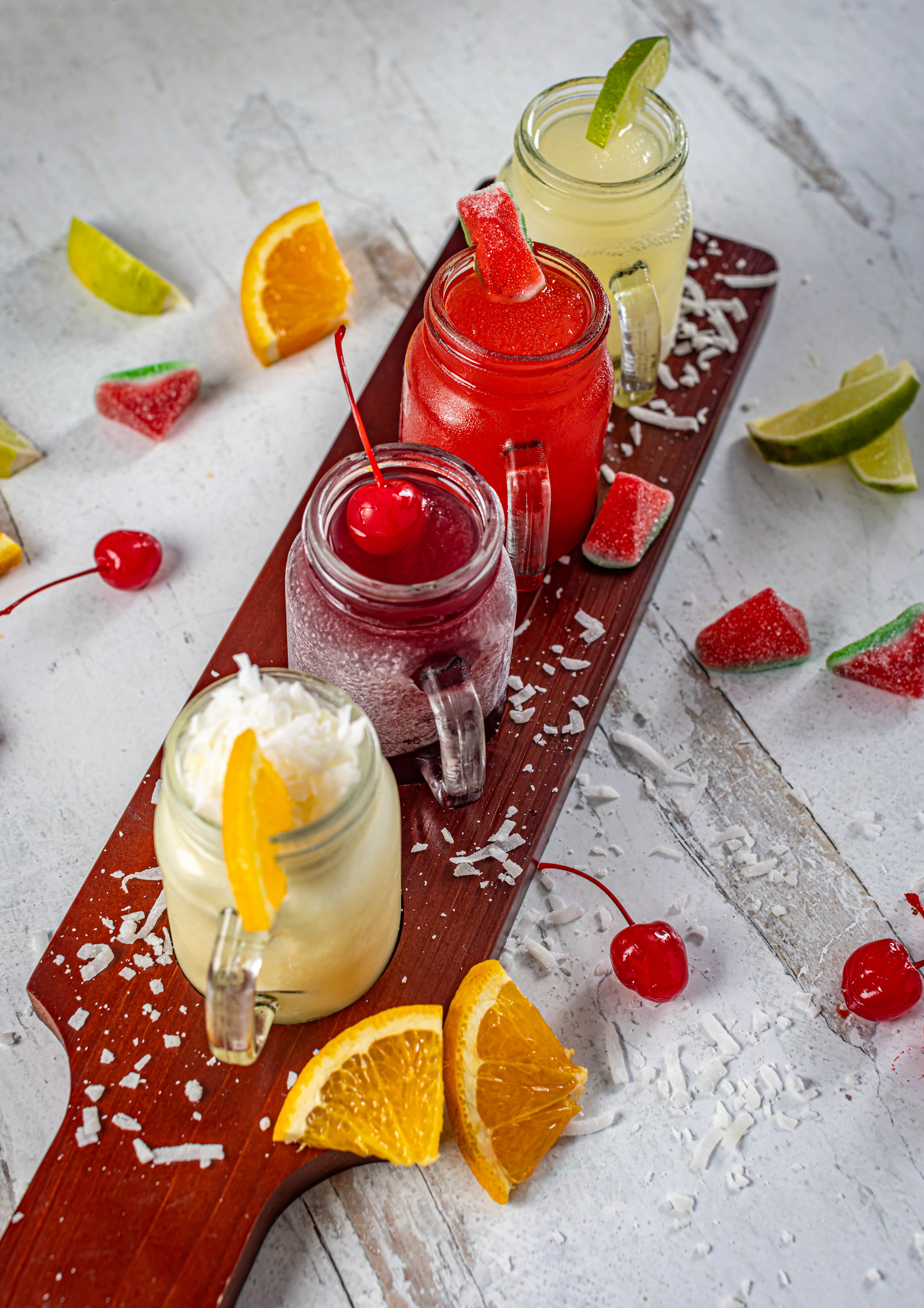 A colorful flight of four refreshing fruit juices, served in mason jar mugs and garnished with fresh fruit and candy.