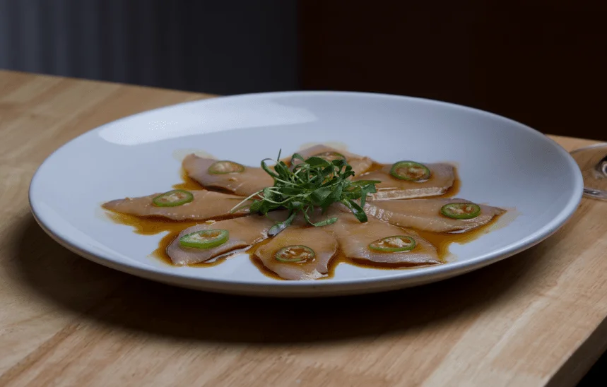 A white round plate featuring yellowtail jalapeño sashimi fanned out in a circular pattern with a light sauce and topped with microgreens, positioned next to a frosted, clear cocktail in a coupe glass garnished with two lychees on a bamboo skewer, all resting on a light wooden table surface.