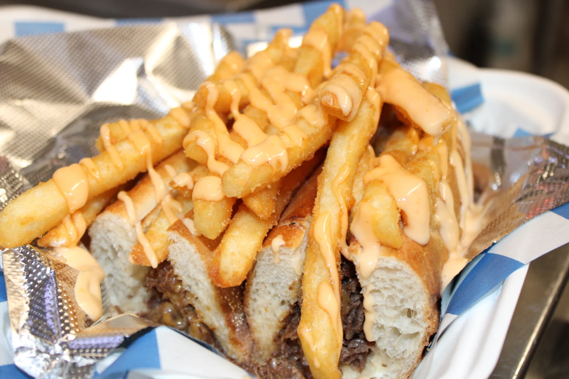 A loaded sandwich topped with crispy fries and drizzled with a creamy sauce, served on a foil-lined tray.