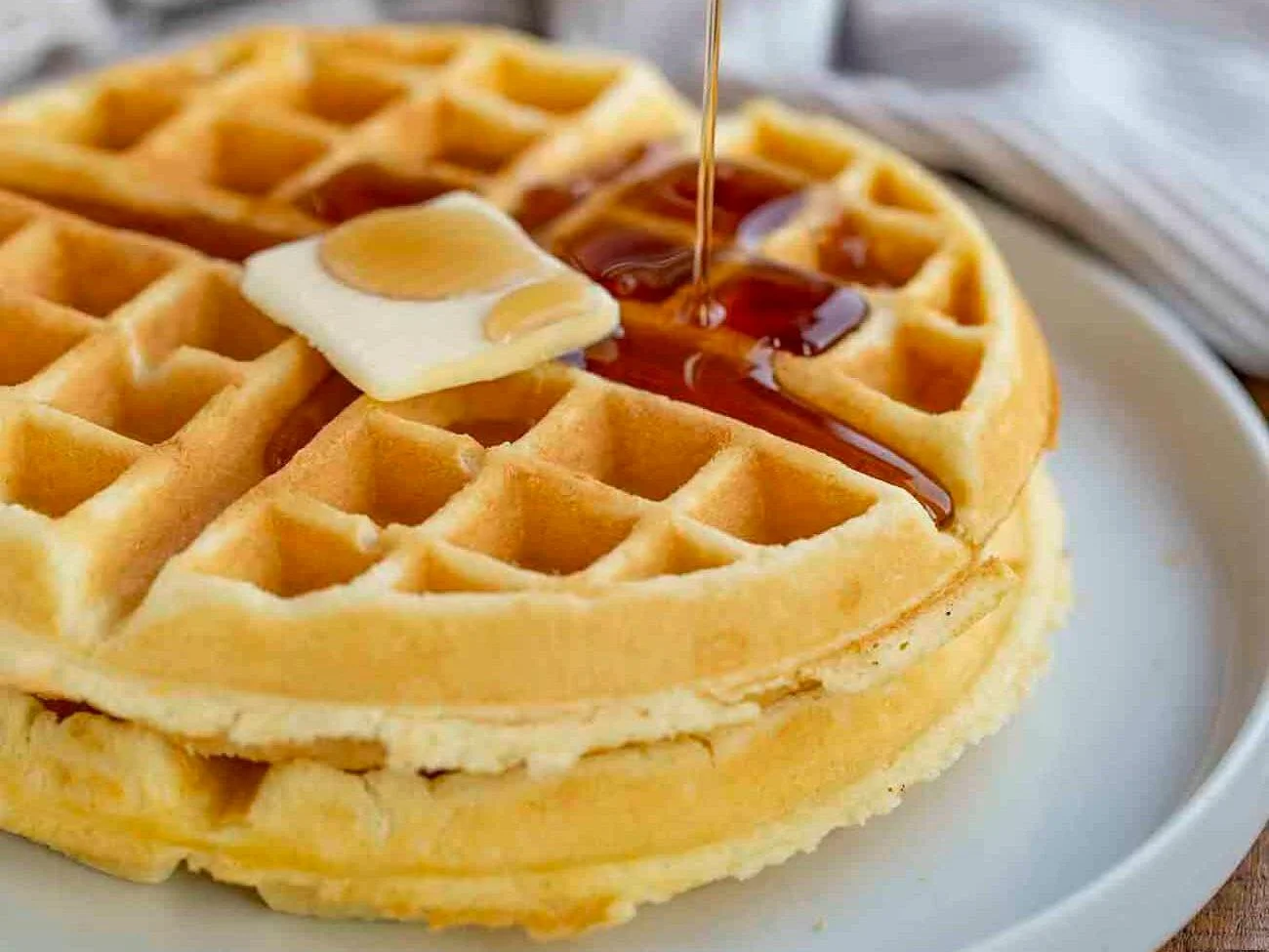 A golden waffle topped with a pat of butter and drizzled with syrup, on a white plate.