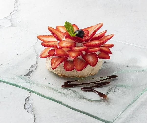A beautifully arranged dessert featuring a layer of whipped cream topped with numerous slices of fresh strawberries, garnished with a mint leaf and a blueberry, presented on a clear glass plate.
