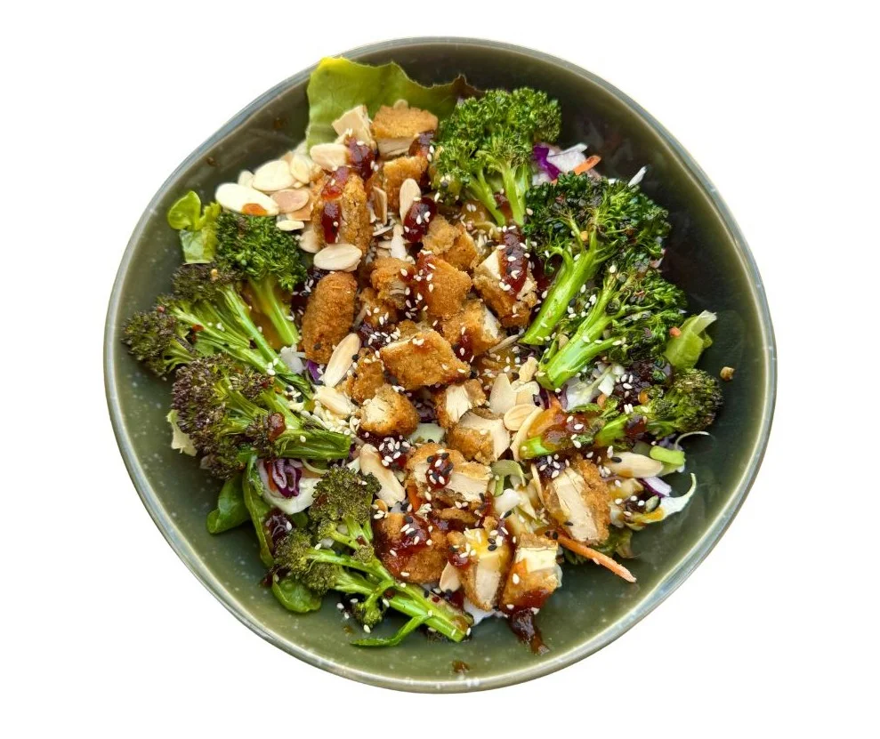 A vibrant salad featuring chopped broccoli, crispy tofu, sliced almonds, and a variety of leafy greens. The dish is topped with a sweet and tangy sauce and sprinkled with sesame seeds.