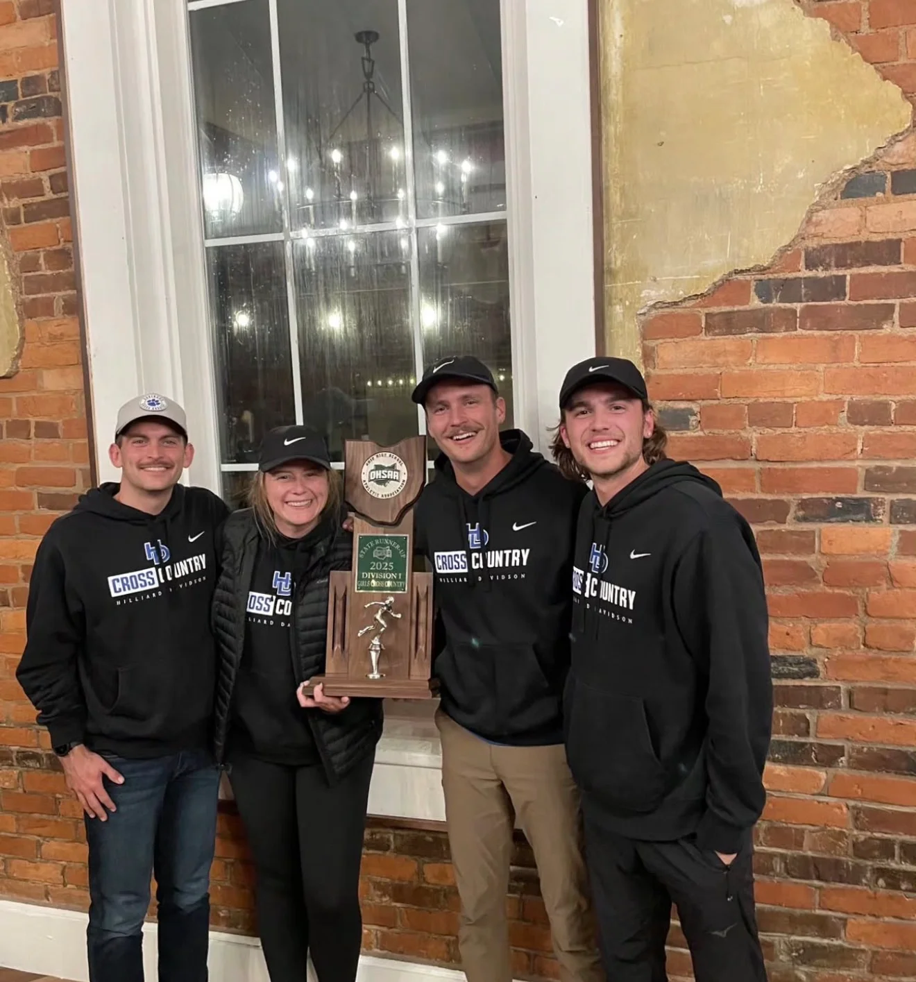 A group of four individuals, dressed in matching black hoodies, celebrating a cross country award, holding a trophy in front of a window with decorative lighting.