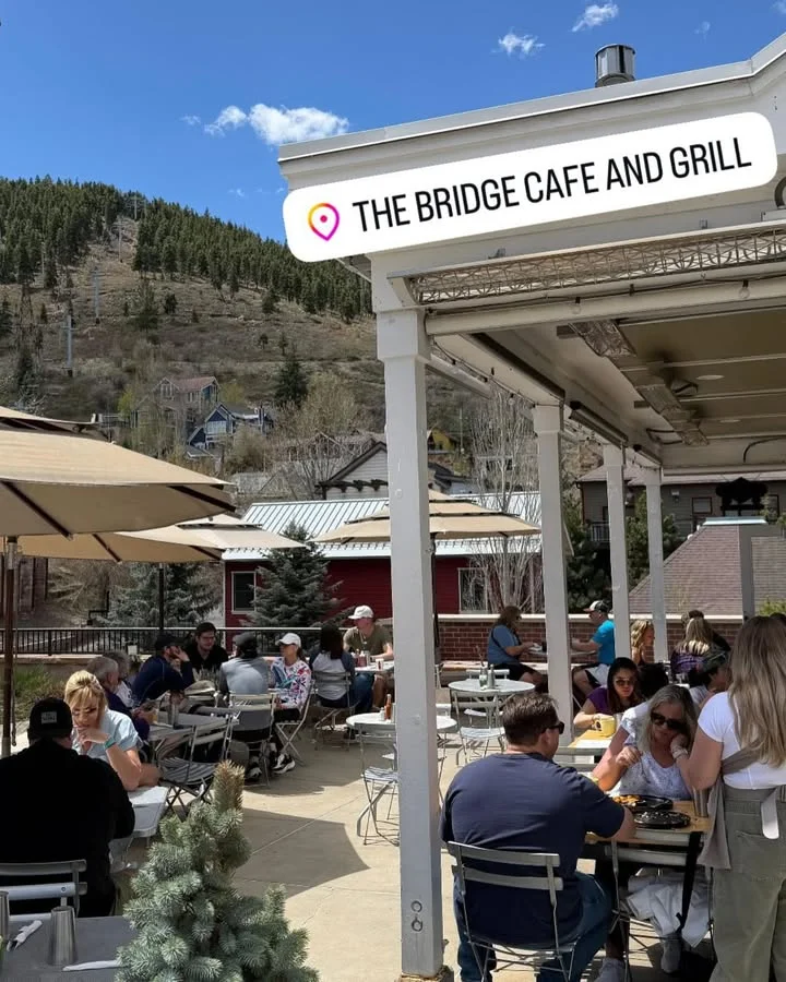An outdoor cafe setting bustling with diners, featuring umbrellas and a scenic view of hills and homes.
