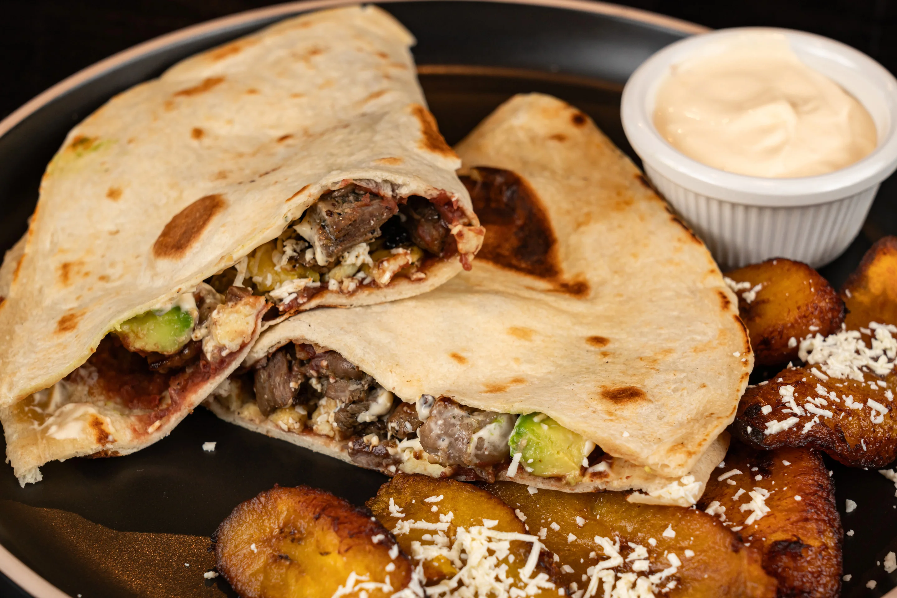 Two folded tortillas filled with meat and toppings, served with a side of sauce and fried plantains.