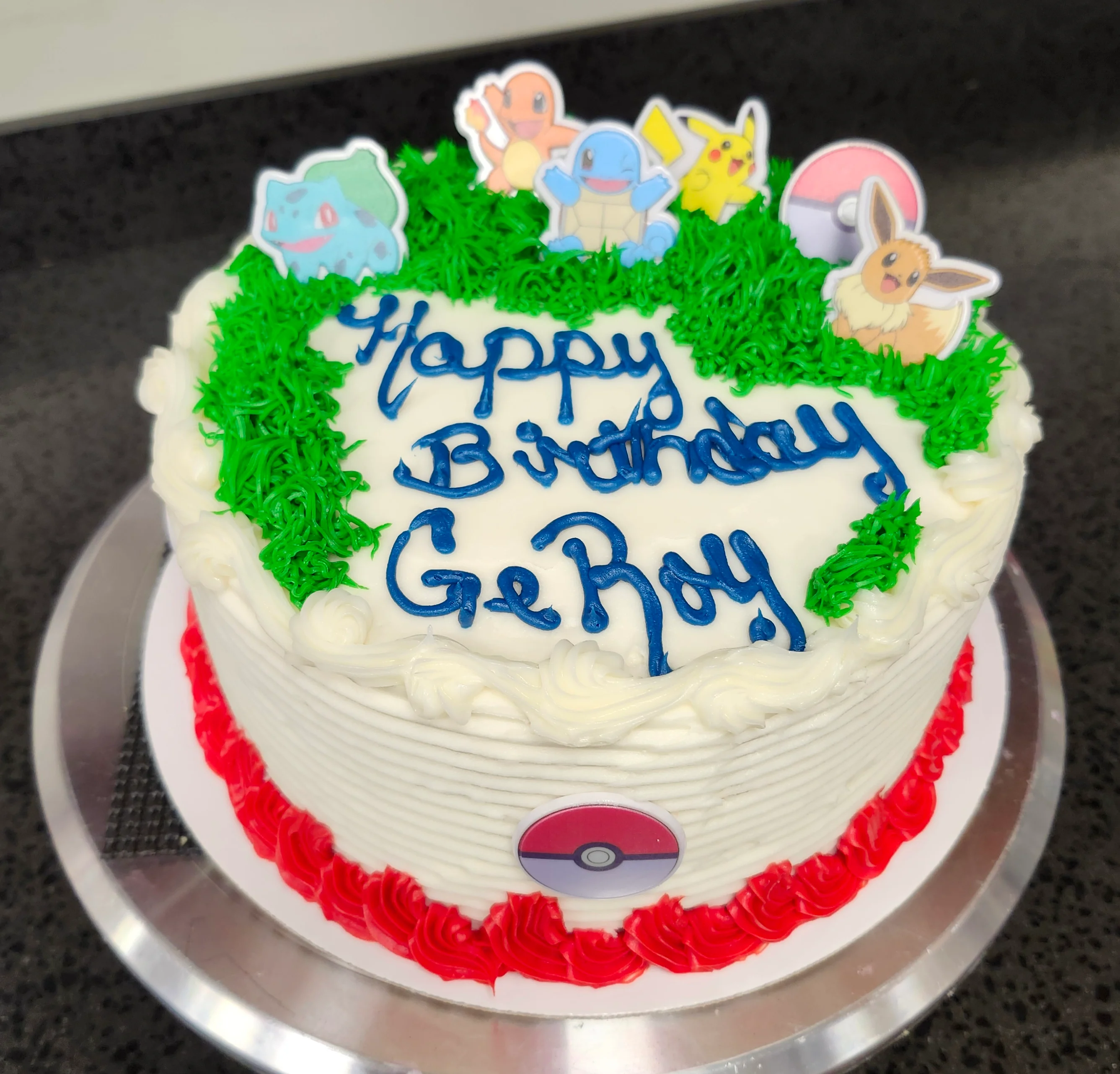 Round cake with decorations of green grass and plastic pokemon that says "Happy Birthday GeRoy"