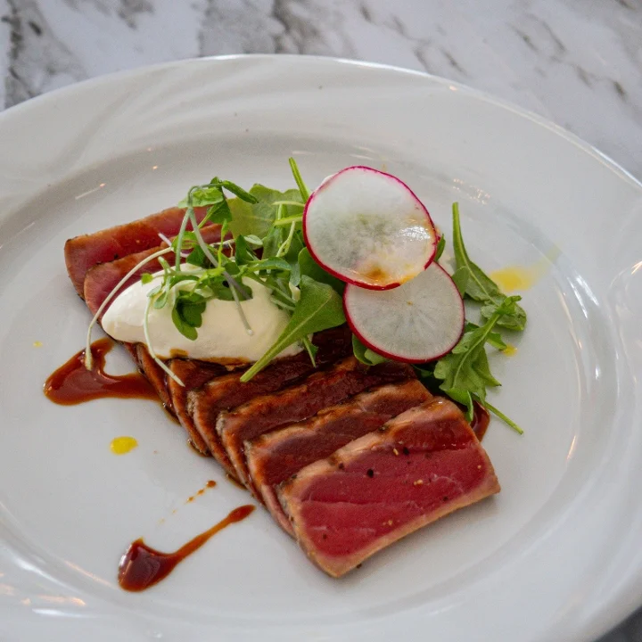 A gourmet dish featuring seared tuna slices, garnished with fresh greens, radish, and a creamy topping on a stylish plate.