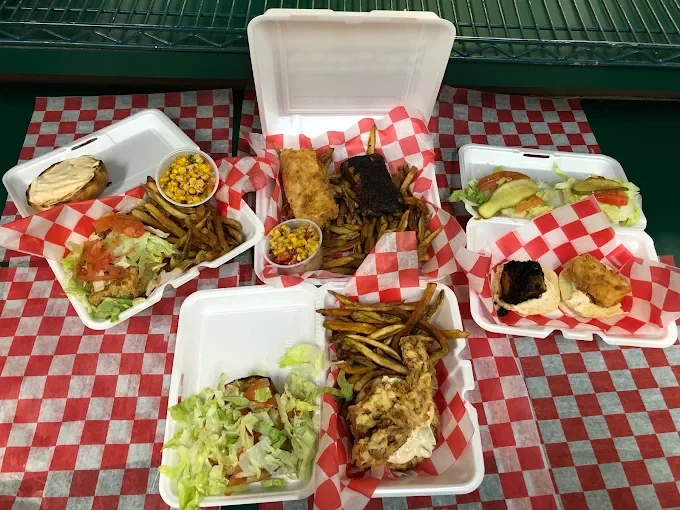 An assortment of takeout meals including grilled chicken, fries, salads, and sandwiches, arranged on a table with red and white checked paper.