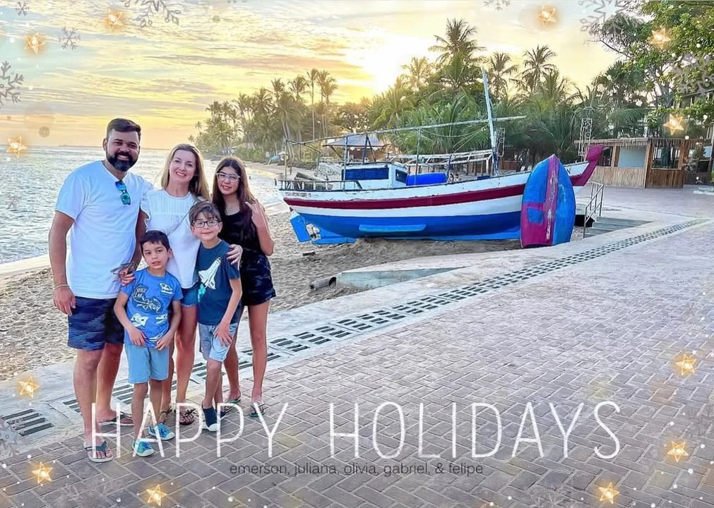 A family posing on a beach at sunset with palm trees and a boat in the background. The scene conveys a tropical holiday atmosphere.