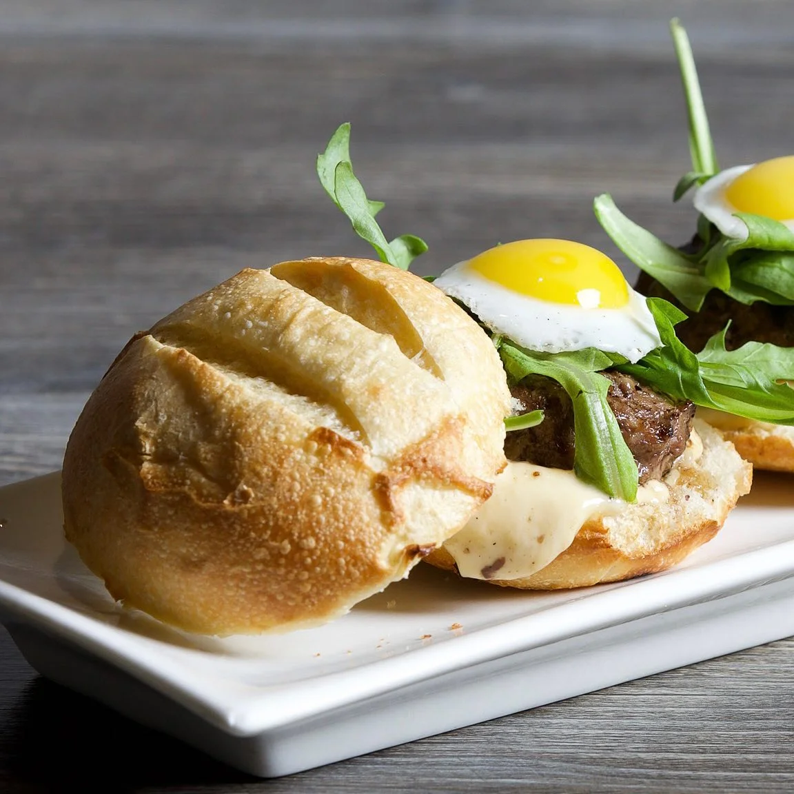 Two mini sliders served on a long white rectangular plate, each featuring a crusty toasted bun, a grilled meat patty topped with melted cheese, fresh arugula leaves, and a sunny-side-up quail egg.