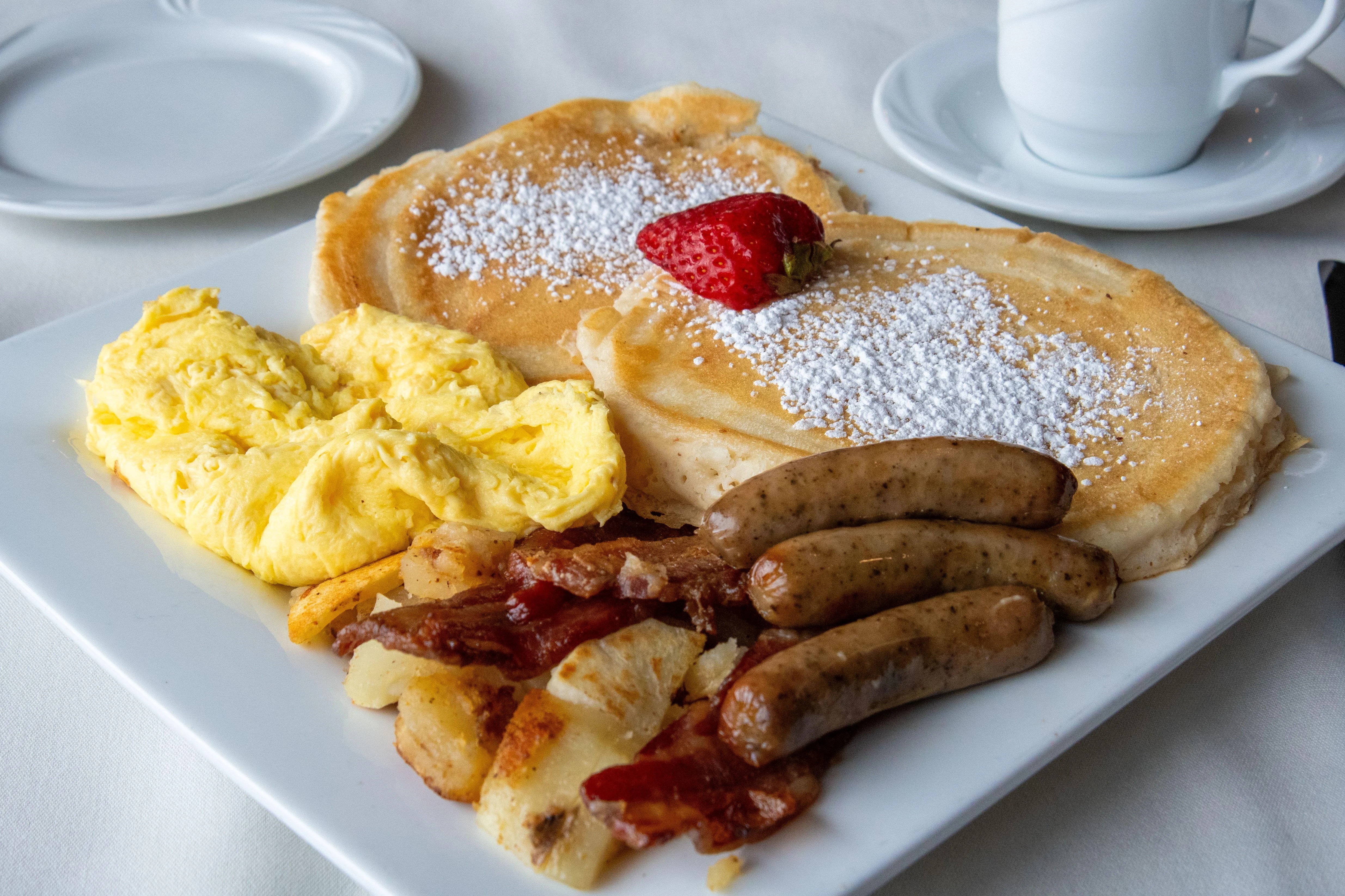 A hearty breakfast plate featuring fluffy pancakes, scrambled eggs, sausages, crispy bacon, and seasoned potato cubes, garnished with a strawberry.