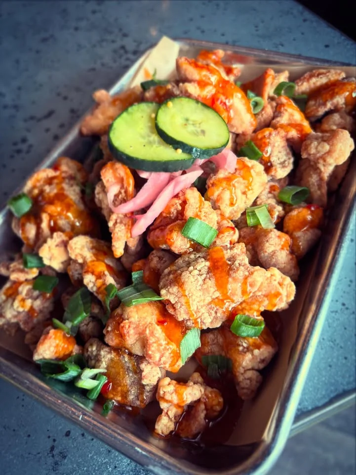 A serving of crispy fried chicken bites topped with sauce, green onions, pickled vegetables, and cucumber slices, presented in a tray.