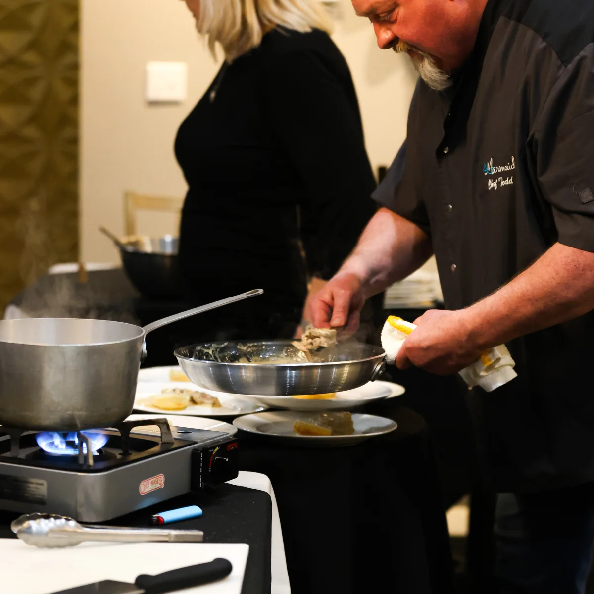 Chef Todd Golden prepares a dish in a stainless steel pan over a portable gas burner, plating food with precision. Another person stands nearby as steam rises from the cooking station.