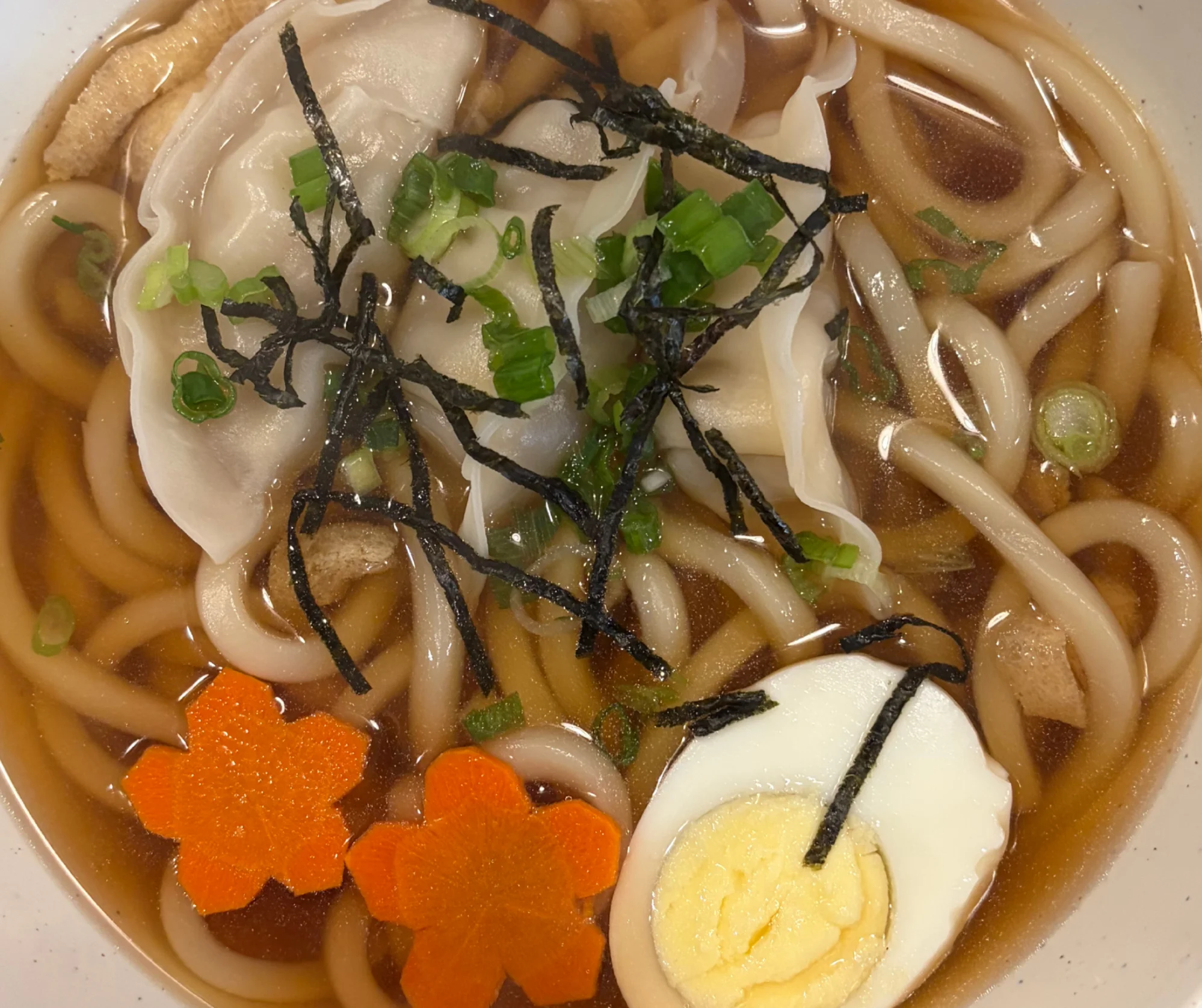 A bowl of noodle soup with udon noodles, soft dumplings, green onions, boiled egg, and decorative carrot flowers, served in a flavorful broth.