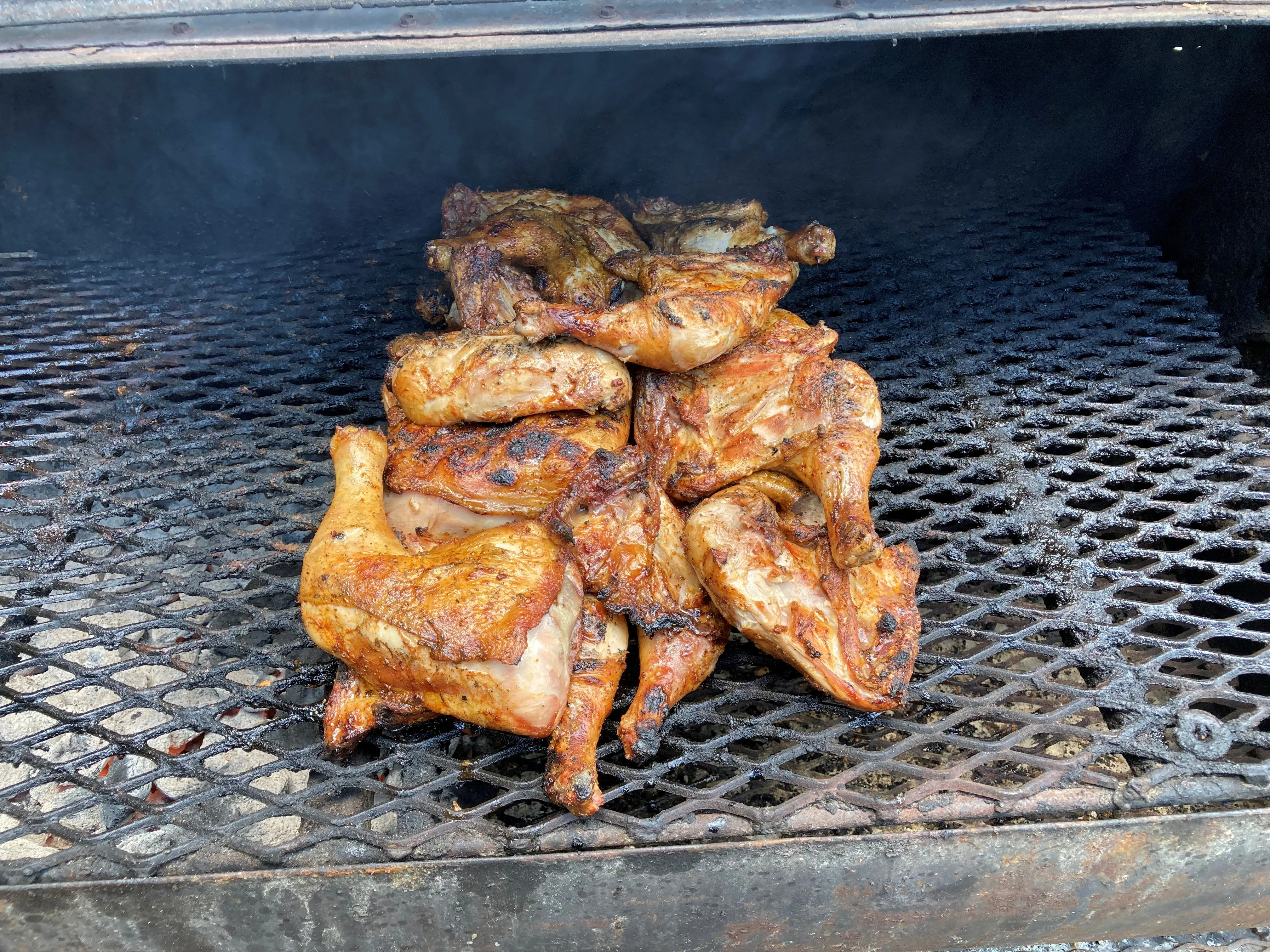 Grilled chicken pieces cooking on a barbecue pit. The chicken is golden brown and appears juicy, resting on a metal grate filled with smoke.