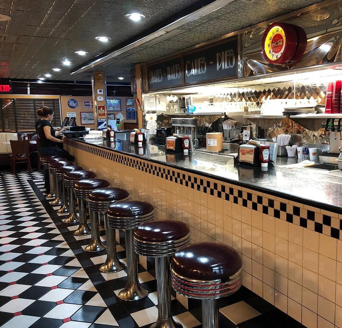 Retro counter seating inside Moonlite Diner featuring classic chrome stools, checkerboard flooring, and an open kitchen serving American comfort food.