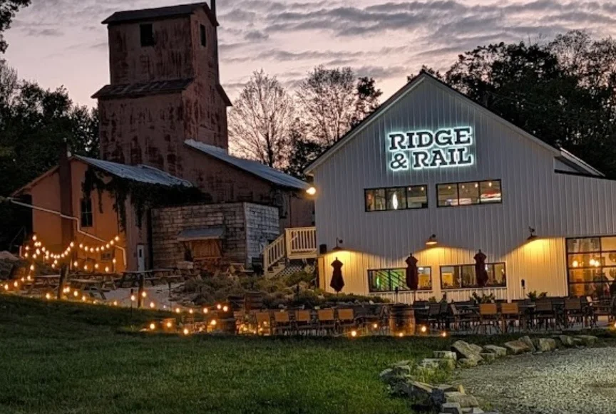 An evening view of a restaurant building with outdoor seating, adorned with string lights. The sky is painted with soft colors of sunset.