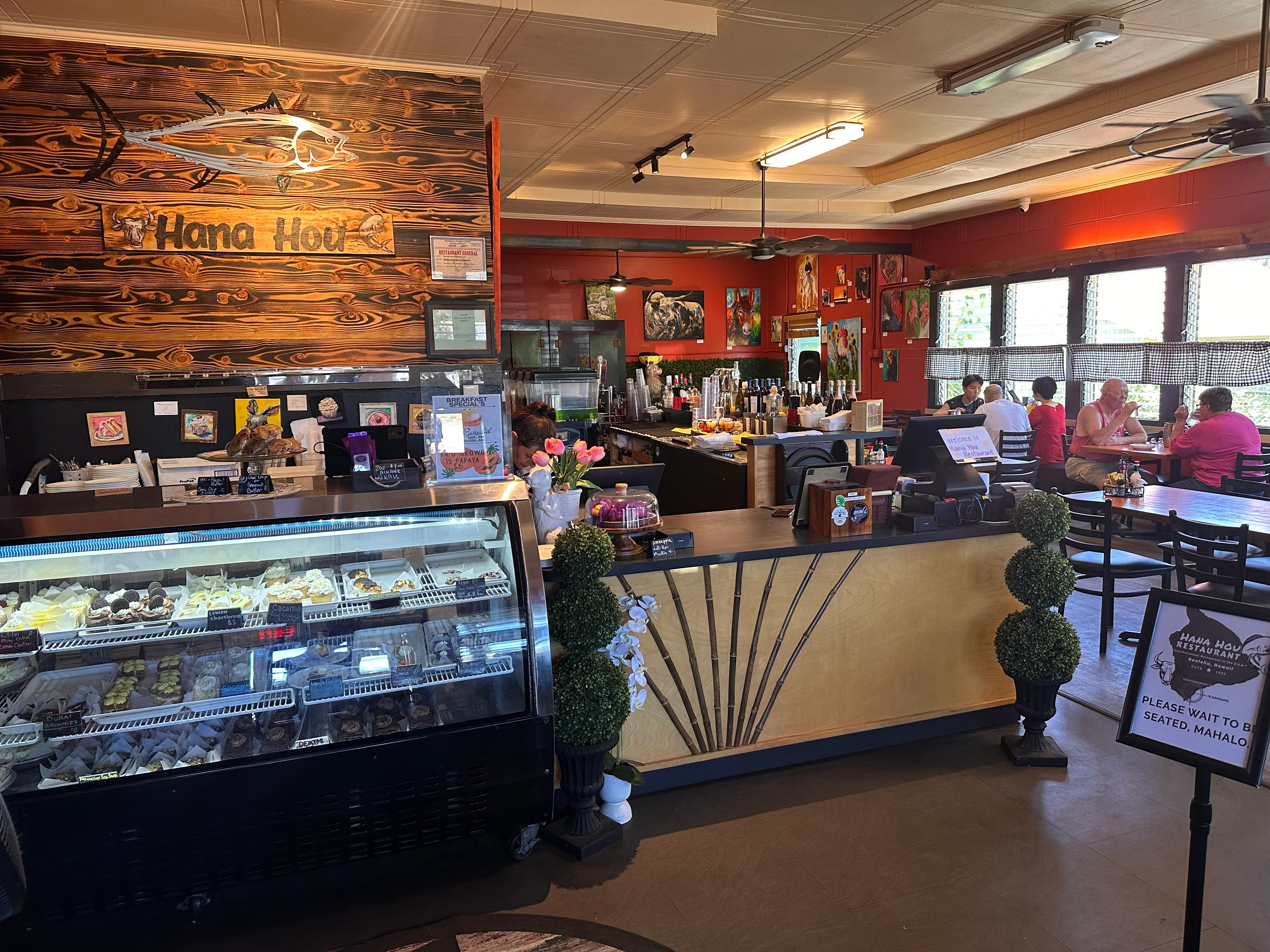An inviting restaurant interior featuring a counter display of desserts, a bar area with drinks, and colorful wall art. Guests are seated at tables enjoying their meals.