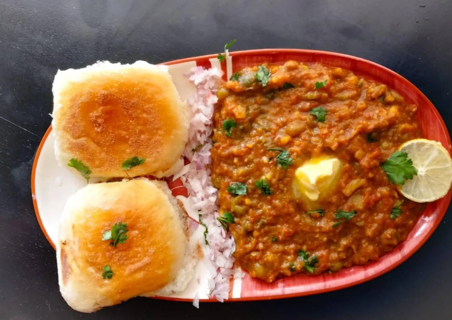 A plate of Pav Bhaji, featuring two buttered buns served with a spicy vegetable curry topped with butter and garnished with cilantro.