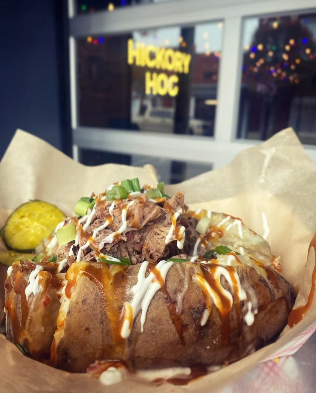 A baked potato topped with shredded meat, green onions, and sauces, served with a pickle on the side.