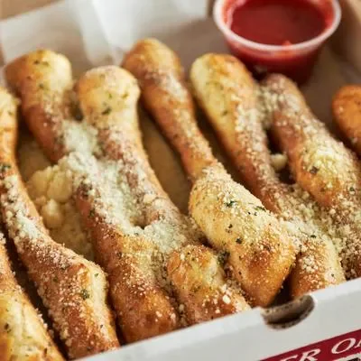 A box containing several breadsticks topped with grated cheese and herbs, accompanied by a small cup of marinara sauce.