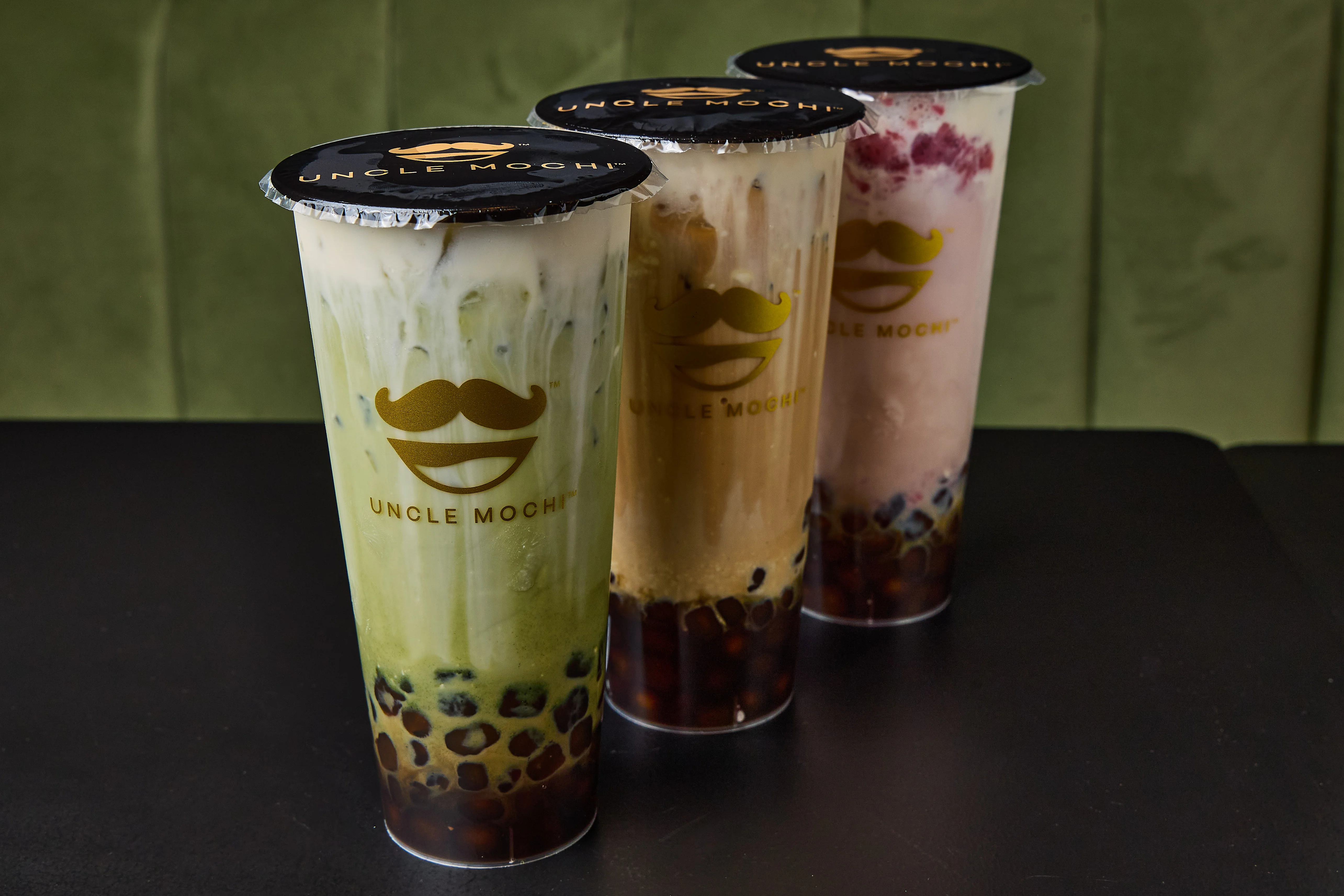 Three colorful bubble tea cups featuring matcha, caramel, and strawberry flavors. Each cup is topped with a distinctive black lid.