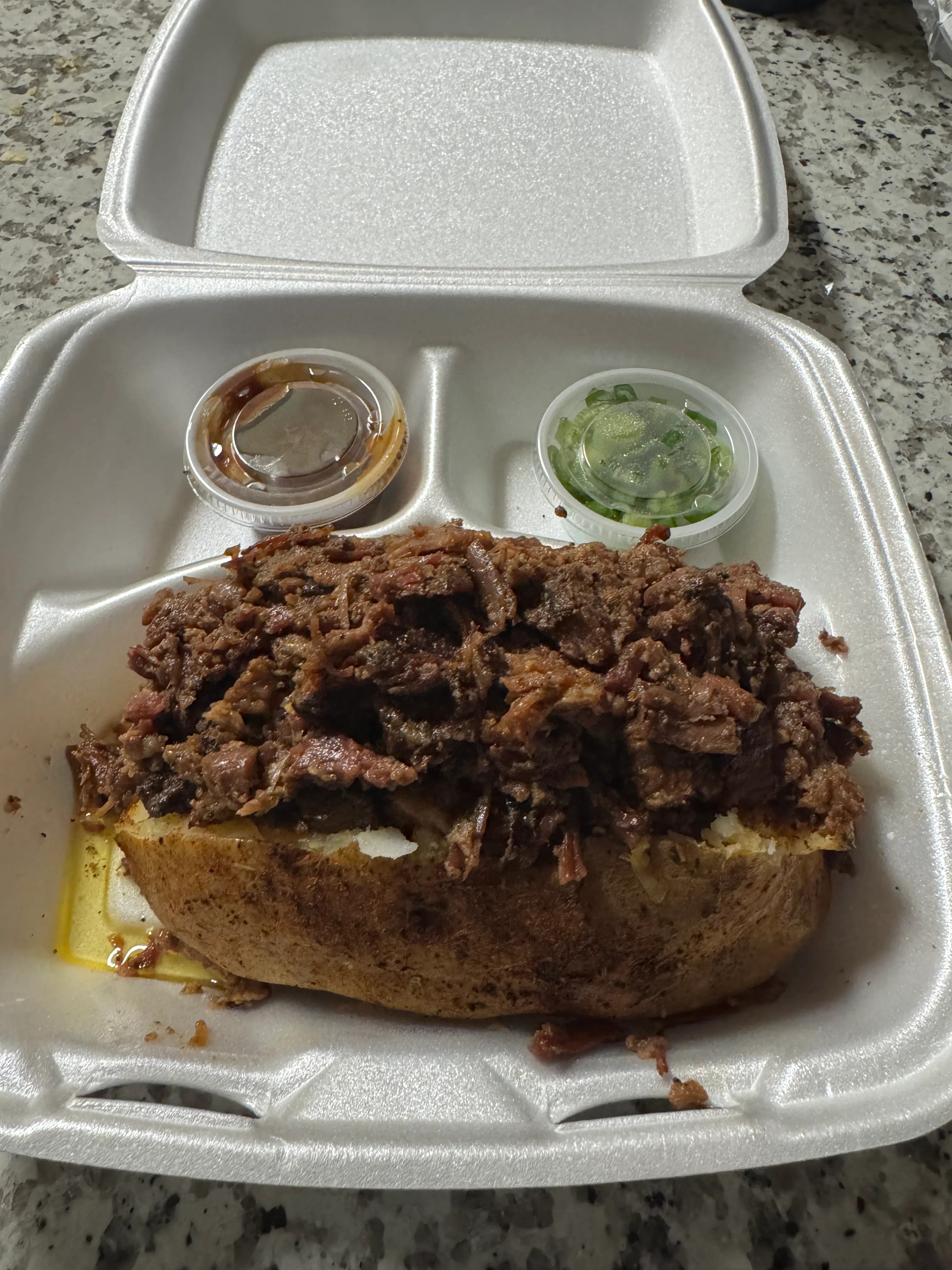 A hearty meat sandwich featuring shredded beef piled on top of a toasted bread, served with two small containers of sauce and chopped green onions.