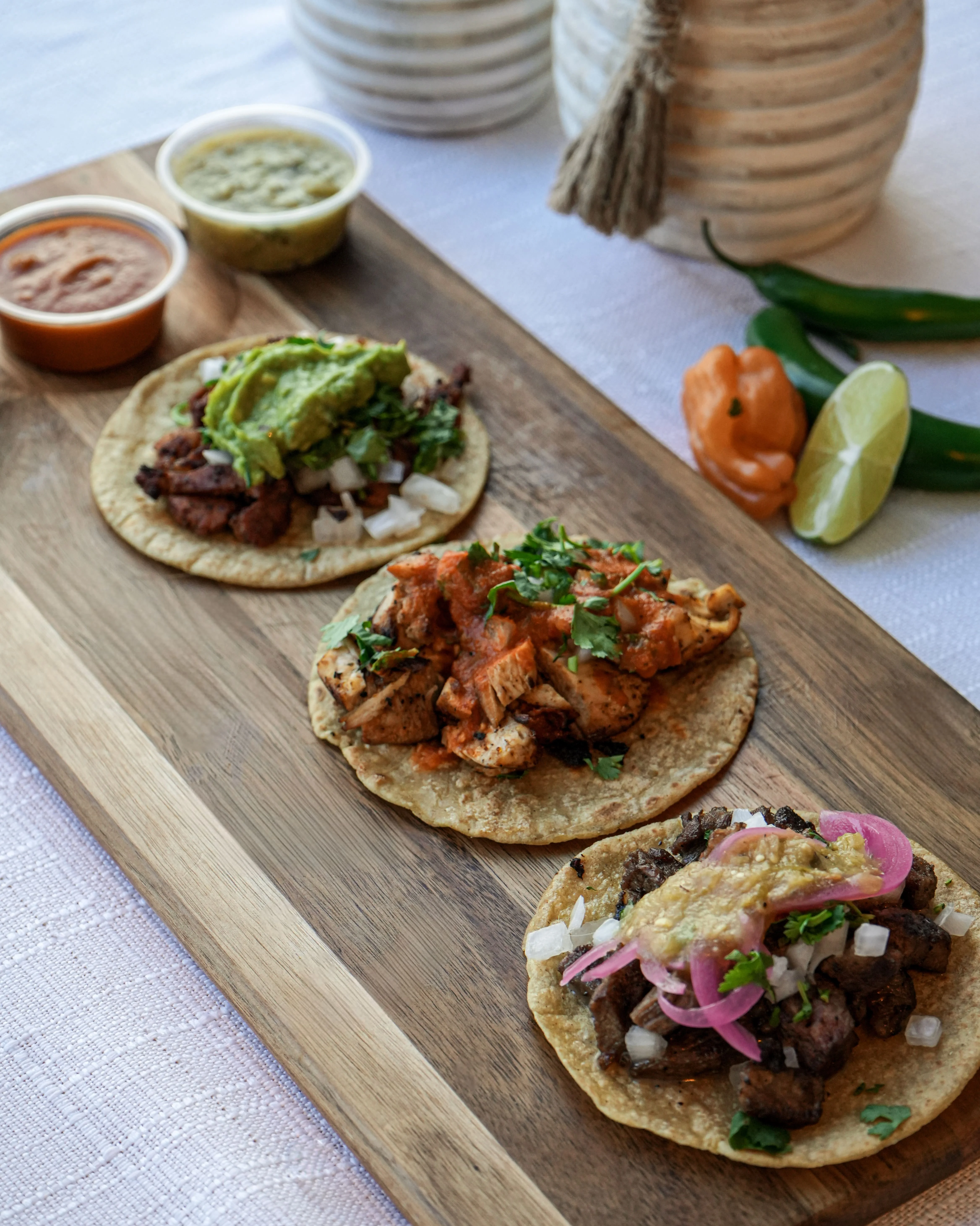 Three tacos on a wooden platter, each topped with different ingredients such as guacamole, meat, and onions, accompanied by colorful sauces on the side.