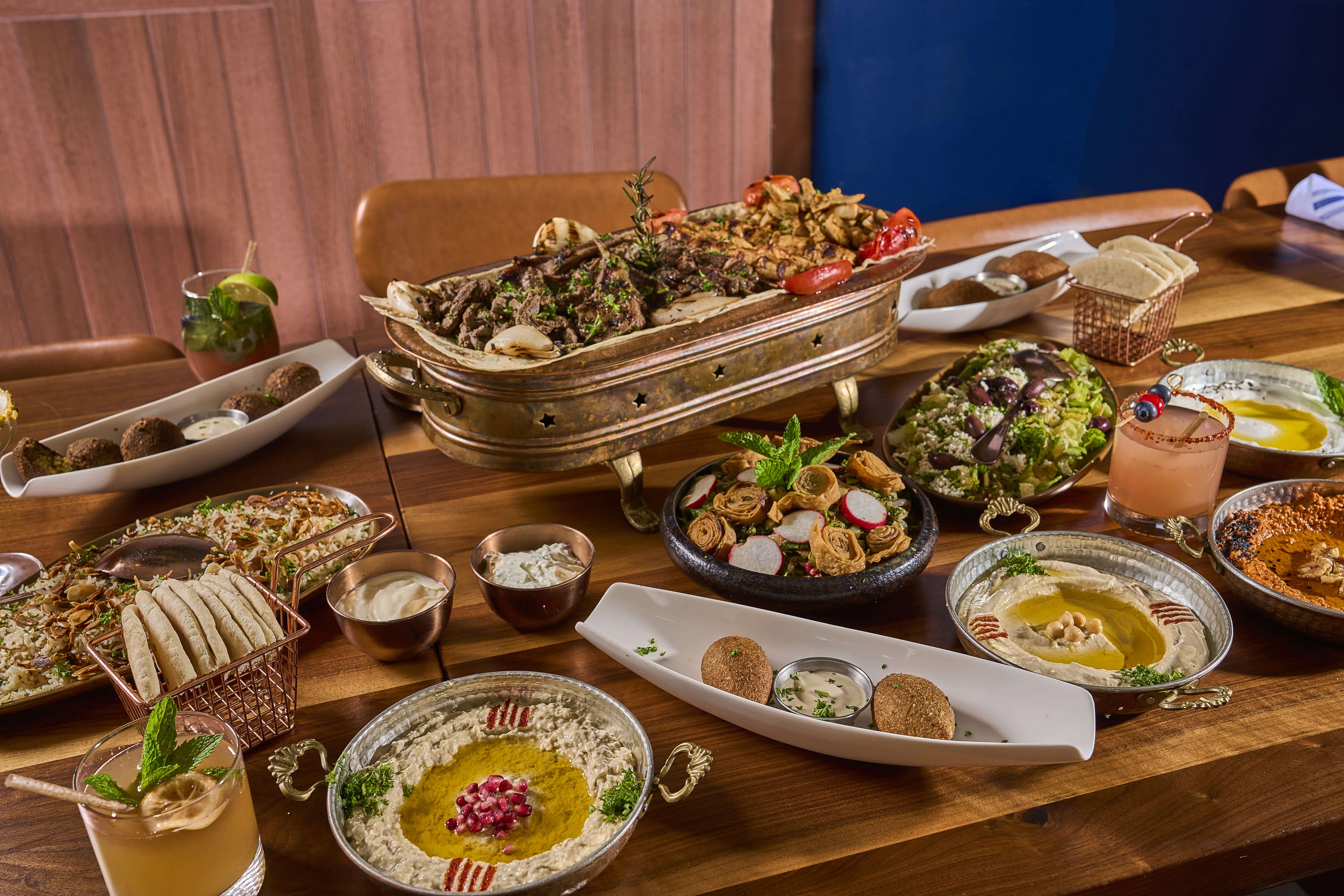 Mediterranean food spread containing grilled meat, salads, falafel, hummus dips, drinks and more. 