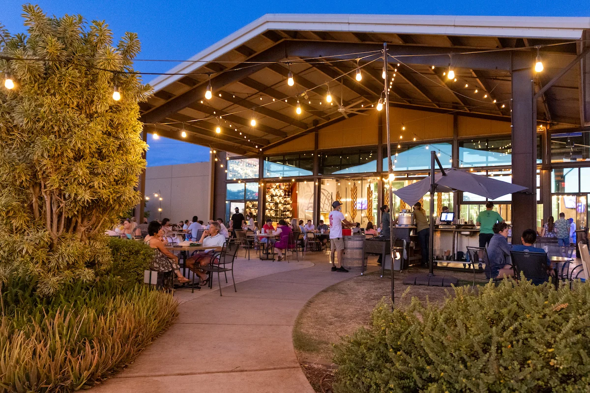 An outdoor dining area at dusk, featuring string lights and an open structure. The space is filled with guests seated at tables, enjoying a warm evening atmosphere.