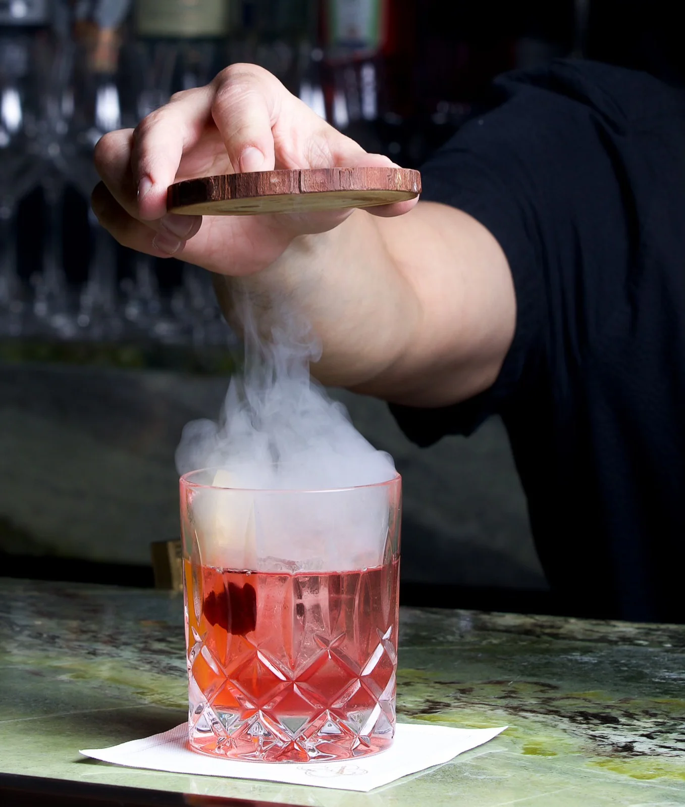 A hand lifting a wooden disc lid from a crystal rocks glass to release a cloud of dense white smoke over a red cocktail with large ice cubes, served on a white napkin over a green marbled bar counter.