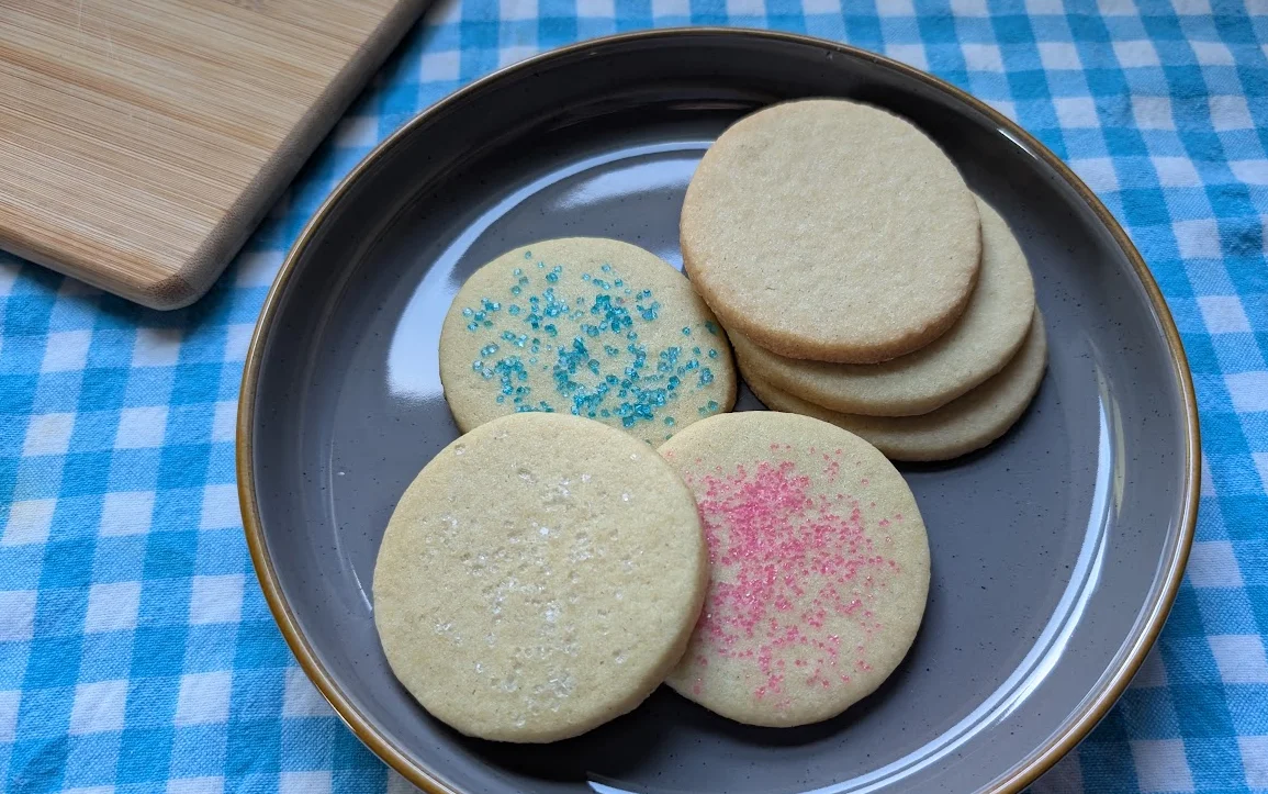 Round sugar cookies with white, blue, and pink sugar sprinkles stacked on a gray plate on a blue gingham tablecloth