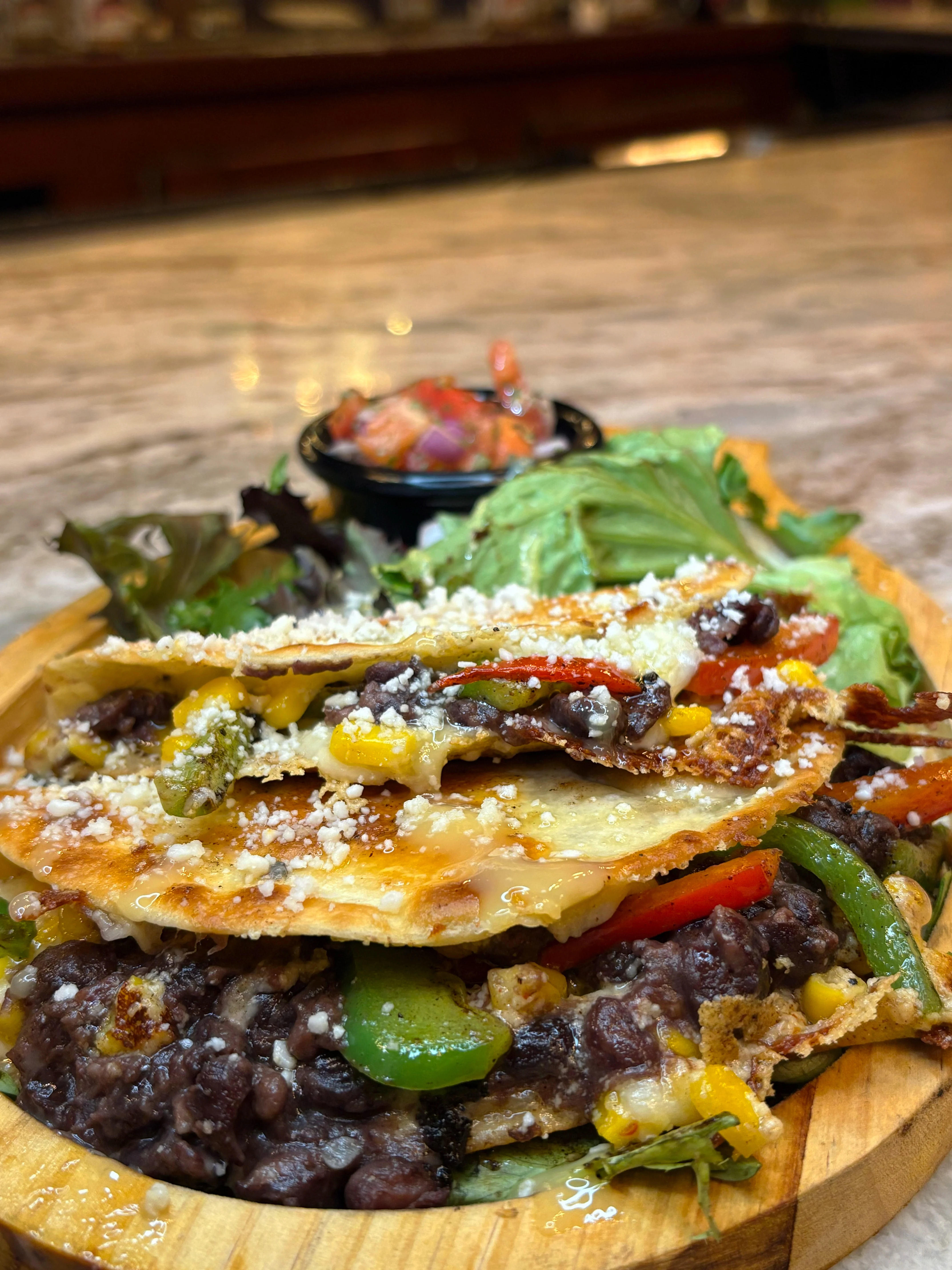 A wooden plate featuring a layered dish of tortillas, black beans, and assorted vegetables topped with cheese, alongside a fresh salsa.
