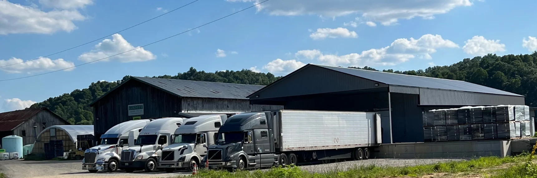 Trucks parked at a rural warehouse under a blue sky with fluffy clouds