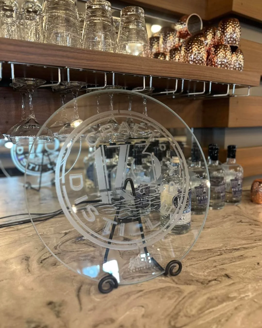 An etched glass sign displaying the name of a craft distillery, accompanied by various glassware on a wooden shelf above a marble surface.