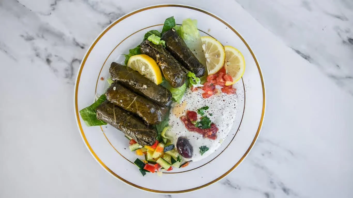 A plate of rolled vine leaves served with lemon slices, fresh vegetables, and a yogurt dip.
